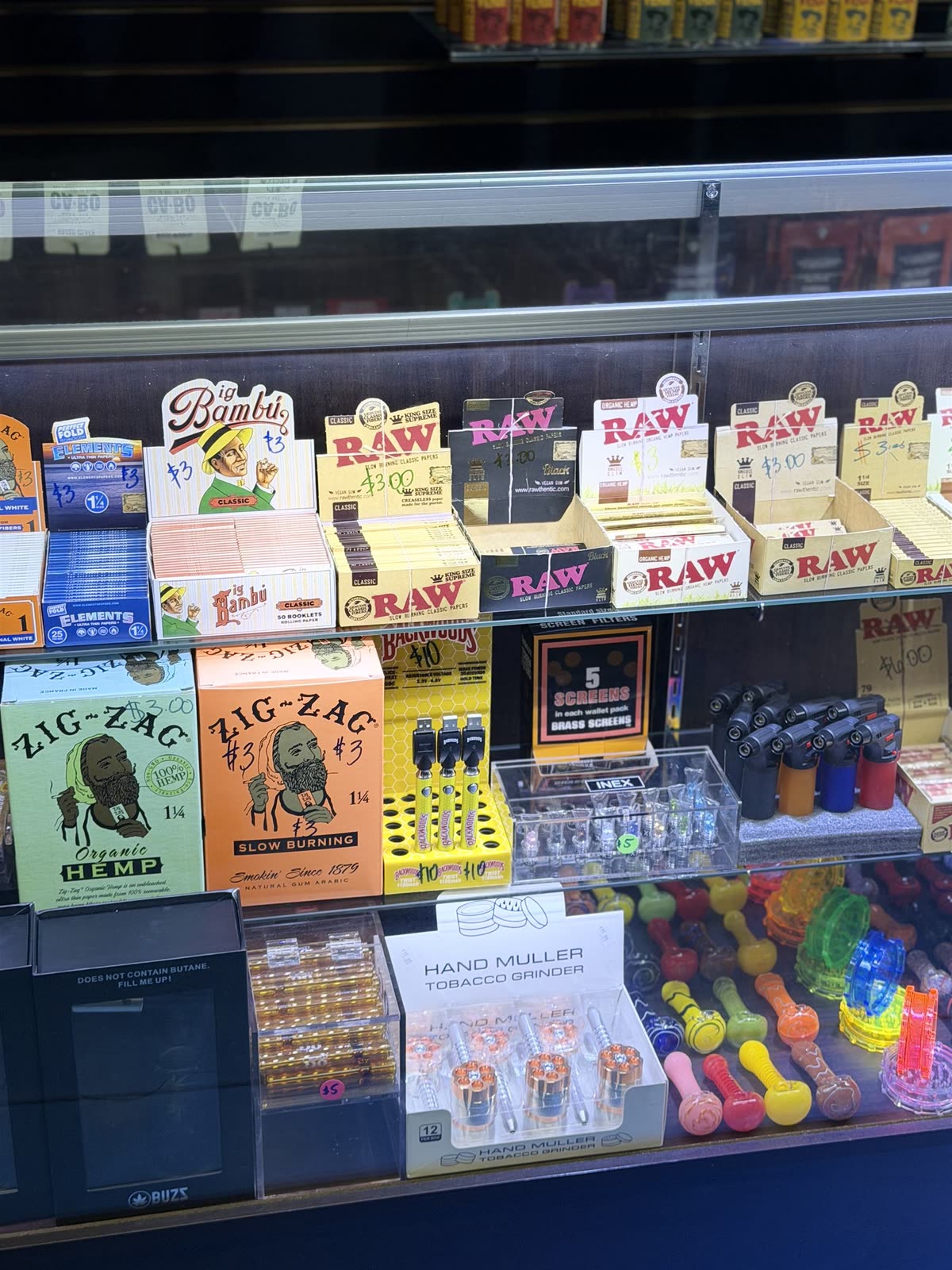 A glass counter case of rolling papers — RAW, Zig-Zag, Elements — with lighters, grinders and colorful hand pipes below.