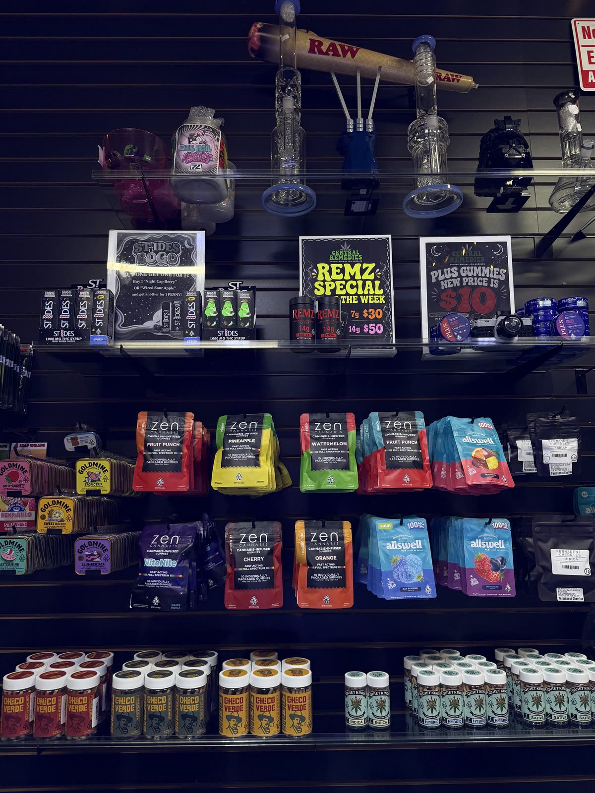 A wall of cannabis-infused gummy pouches in bright colors, with hand-lettered deal signs above the shelf.