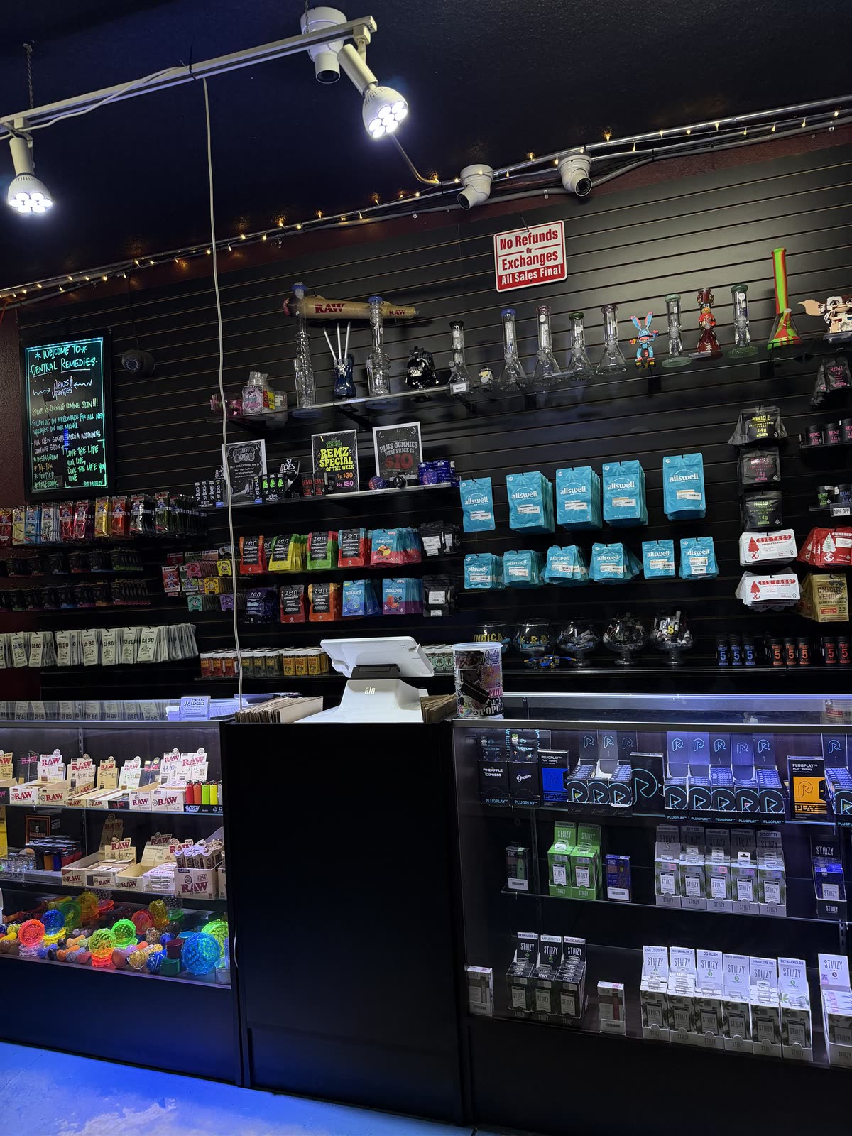 A neon welcome board reading 'Welcome to Central Remedies' beside racks of wraps and vape products on black slat walls.